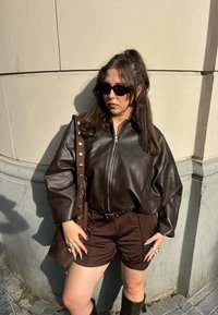 Barna bőr cipzáras dzseki túlméretezett ujjal, barna rövidnadrággal és hozzáillő kézitáskával párosítva. Kiegészítők: napszemüveg és gyűrűk.