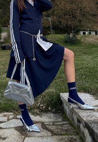 Personne en tailleur jupe bleu marine avec rayures blanches, ceinture chaîne argentée, sac à main métallique, chaussettes Adidas bleu marine et chaussures à talons hauts argentées en extérieur.