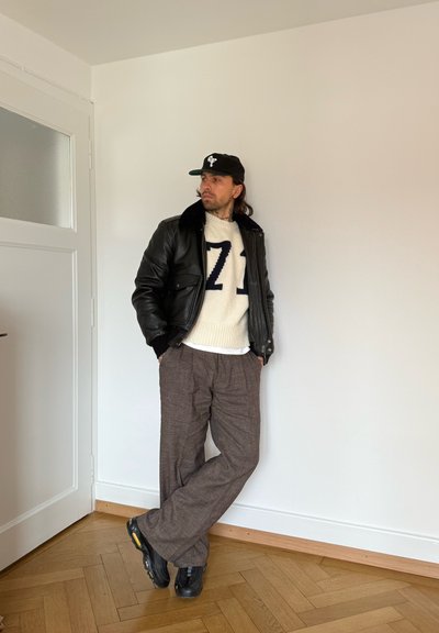 Hombre con gorra negra, chaqueta de cuero negra, suéter blanco con "71", pantalones marrones y zapatillas negras, apoyado contra una pared blanca en el interior.