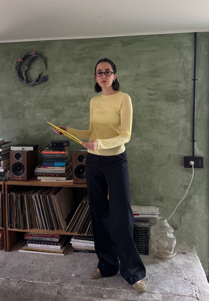 Camisola amarela de mangas longas com riscas, calças largas pretas com riscas, segurando um papel amarelo, em pé ao lado de uma prateleira de madeira com discos de vinil.