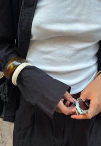 Persoon in een zwart shirt met lange mouwen over een wit geribbeld topje, met in de hand een zonnebril en twee stevige armbanden om de linkerpols.