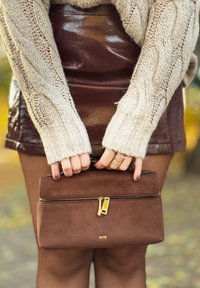 Braune Wildleder-Clutch mit goldenem Reißverschluss und Beschlägen, mit beiden Händen gehalten, kombiniert mit einem cremefarbenen Strickpullover und einem glänzenden burgunderroten Rock.