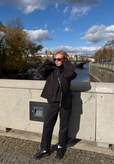 Chaqueta negra de gran tamaño y pantalones a juego, combinados con zapatos negros. Accesorios incluyen gafas de sol y un pequeño bolso bandolera. Entorno urbano.