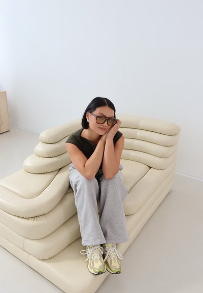 Una joven con el cabello corto y negro y gafas de sol está sentada, sonriendo, en un sofá moderno beige en una habitación minimalista blanca.