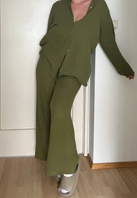 Camisa verde-oliva, texturizada, de botões, combinada com calças largas na mesma cor; pessoa a usar pantufas bege e meias brancas.