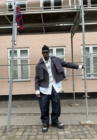 Lyhyt harmaa takki valkoisen paidan päällä, yhdistettynä väljiiin tummansinisiin farkkuihin ja järeisiin mustiin kenkiin. Henkilöllä on aurinkolasit ja pipo.