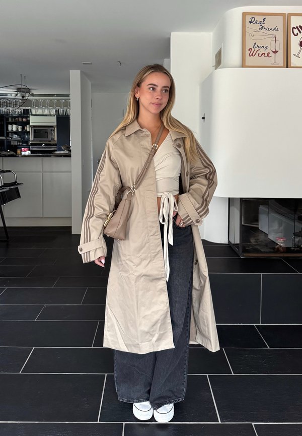 Joven mujer con abrigo beige tipo gabardina, camiseta blanca con nudo delante, pantalones anchos negros y zapatillas blancas, de pie en el interior junto a una chimenea y cocina modernas.