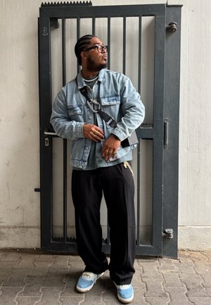 Chaqueta de mezclilla sobre una camiseta verde, pantalones anchos negros y zapatillas azules. Los accesorios incluyen anillos y un bolso cruzado. Puerta gris en el fondo.