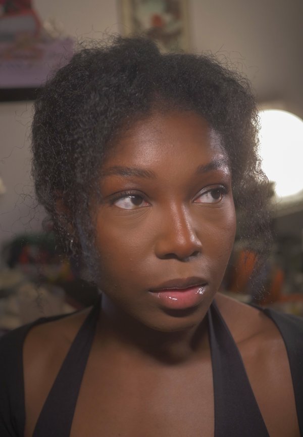 Femme aux cheveux bouclés et lèvres brillantes regardant légèrement vers la droite, avec un arrière-plan doucement flou et un éclairage chaud.