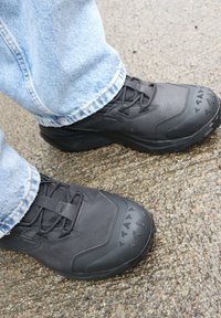 Chaussures de sécurité noires à lacets avec embouts texturés portées avec un jean bleu clair, debout sur un sol en béton rugueux.