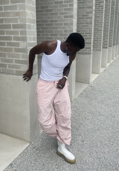 Top blanco sin mangas, pantalones sueltos rosa claro y botas blancas hasta el tobillo con una suela gruesa de color verde claro. El fondo presenta una pared de ladrillos gris.