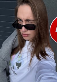 White long-sleeve shirt with colorful abstract design, gray oversized jacket, and black oval sunglasses. Straight brown hair. Backdrop is gray.