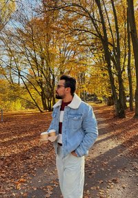Mann i denimjakke som holder to takeaway-kaffer, stående på en løvdekket sti omgitt av høsttrær i en park.