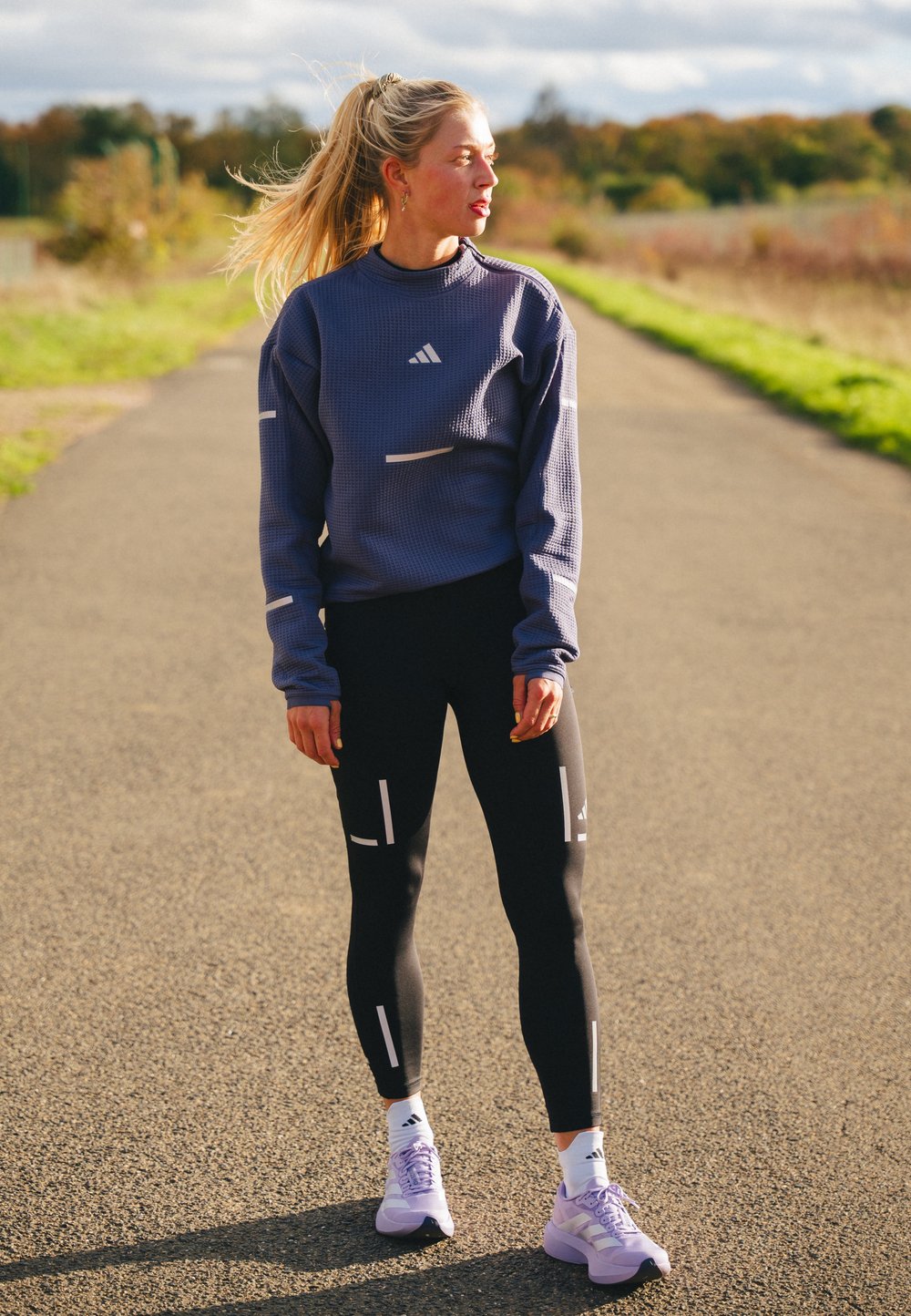 Paarse sweater met wafelstructuur en witte accenten, zwarte legging met verticale prints, en paarse sportschoenen met witte details.