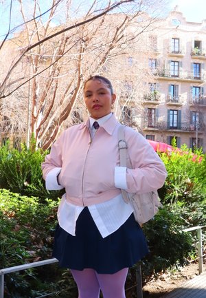 Mujer joven que lleva una chaqueta rosa, camisa blanca, falda azul marino y medias lavanda, de pie al aire libre con edificios urbanos y árboles sin hojas detrás de ella.