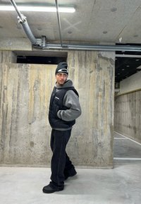 Uomo con berretto nero, gilet imbottito nero, felpa grigia con cappuccio e pantaloni neri in piedi in un parcheggio industriale in cemento, mentre guarda di lato.