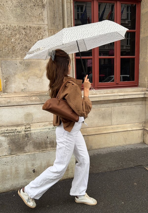 Femme marchant sur un trottoir mouillé, tenant un parapluie blanc à pois noirs, portant une veste marron, un pantalon blanc, et portant un sac à main marron.