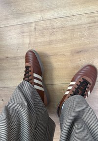 Brązowe skórzane buty z białymi paskami, teksturowanym wzorem, czarne sznurowadła, zestawione z szarymi spodniami w kratę na drewnianej podłodze.