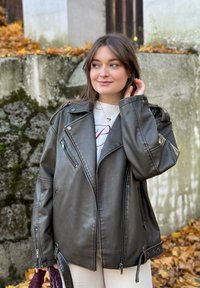 Veste de moto en simili cuir noir avec quincaillerie argentée, fermetures éclair et large col. Elle possède des épaules tombantes et deux poches latérales.