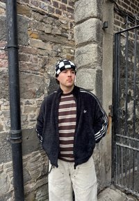 Jeune homme portant un bonnet à carreaux, une chemise rayée, une veste noire duveteuse et un pantalon cargo clair, debout contre un mur en pierre près d'une grille métallique.