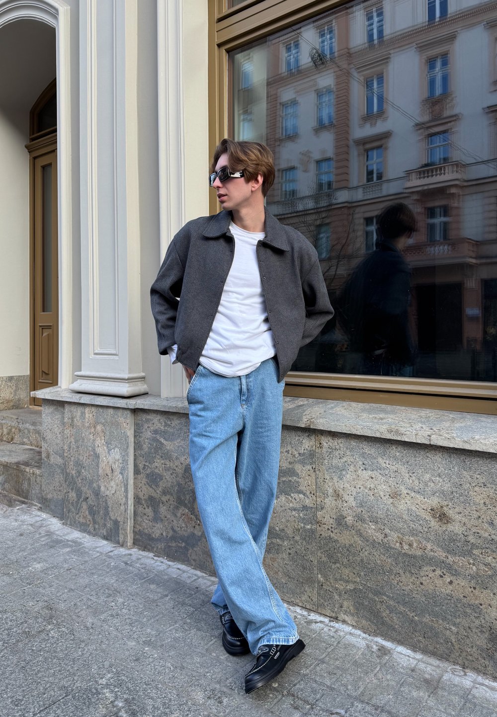 Graue Cordjacke, weißes T-Shirt, hellblaue weit geschnittene Jeans und schwarze Schuhe. Steht an einer Steinewand mit einem reflektierenden Fenster im Hintergrund.
