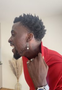 Orecchino a cerchio d'argento con una perla, camicia rossa con colletto, capelli corti e texturizzati, anelli d'argento e un orologio, che raffigura una vista laterale.