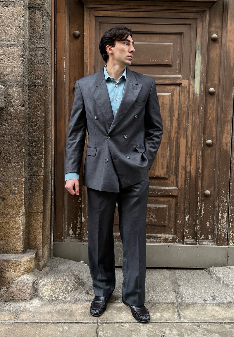 Homme en costume gris foncé à double boutonnage et chemise bleu clair, debout avec une main dans la poche, regardant vers sa gauche devant une porte en bois.