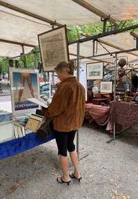 En kvinne iført en brun semsket skinnjakke og svarte leggings undersøker en bok på et marked, omkranset av vintageartikler og kunstverk.