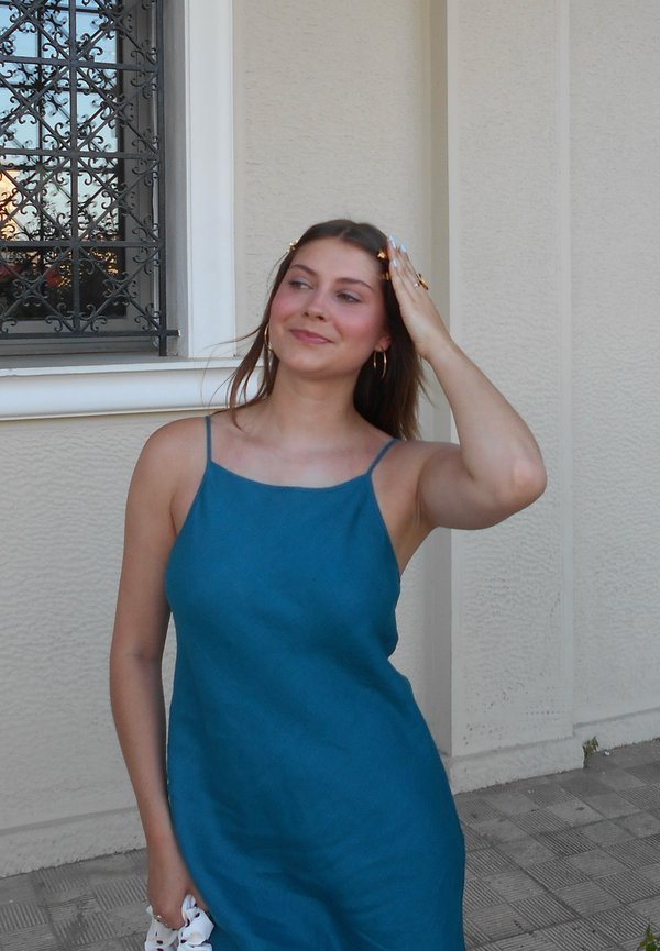 Junge Frau in blauem ärmellosem Kleid berührt ihr Haar und steht draußen neben einer beige Wand und einem Fenster mit schwarzen Metallstäben.