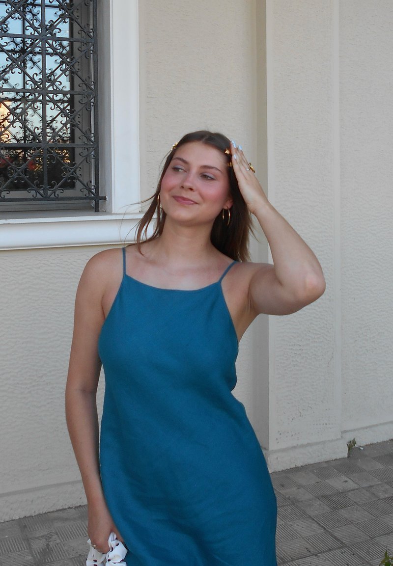 Jeune femme en robe bleue sans manches touchant ses cheveux, debout à l'extérieur près d'un mur beige et une fenêtre avec des barreaux métalliques noirs.