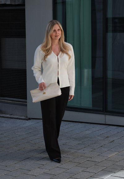 Mujer con cabello largo y rubio, vestida con blusa blanca y pantalones negros, sosteniendo un bolso de mano beige, de pie en una zona pavimentada cerca de un edificio de cristal.