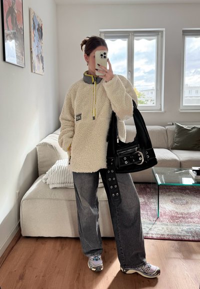 Suéter de forro polar en color blanco roto con cuello gris y cremallera amarilla. Jeans grises sueltos con hebillas laterales, bolso negro y zapatillas multicolor.