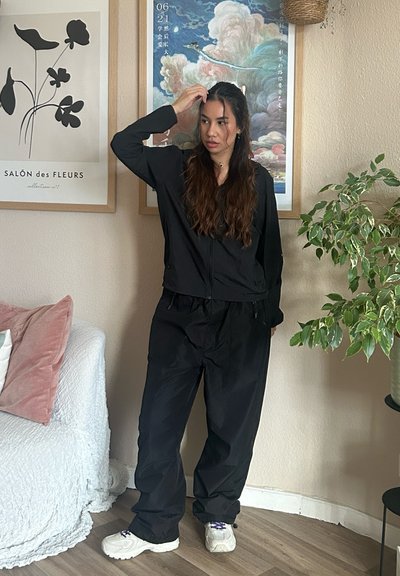 Chaqueta negra con cremallera y mangas largas, combinada con pantalones negros holgados. Zapatillas blancas resistentes. Ambiente interior con plantas y arte enmarcado.