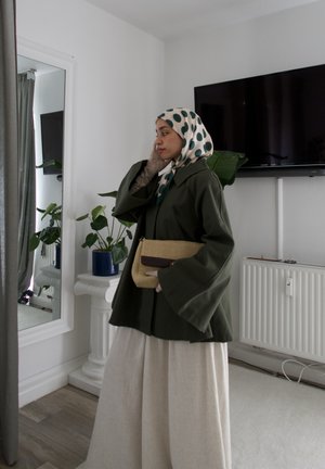 Abrigo verde oliva con mangas anchas, pantalones fluidos en tono crema y un bolso beige con un acento marrón, combinado con un pañuelo de cabeza estampado.