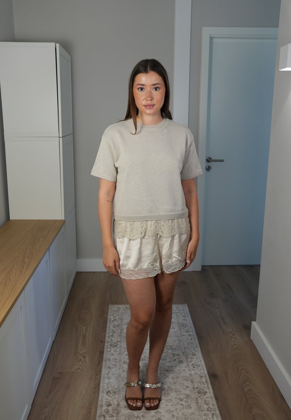 Jeune femme debout à l'intérieur sur un tapis, portant un pull beige à manches courtes, un short en dentelle et des sandales ouvertes.