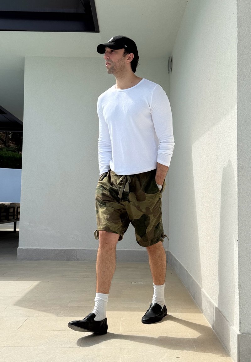 Homme marchant à l'intérieur portant une chemise blanche à manches longues, un short camouflage, des chaussettes blanches, des chaussures noires et une casquette noire.