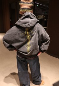 Felpa grigia con cappuccio e motivo di rametto giallo sul retro, abbinata a pantaloni di denim scuro e larghi. La texture appare morbida e casual.