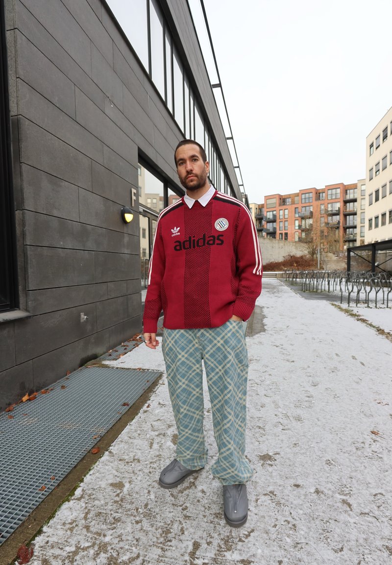 Pull en maille rouge avec un col blanc et un logo "adidas" noir, associé à un pantalon à carreaux bleu clair et des bottes grises, posé en plein air sur un chemin enneigé.