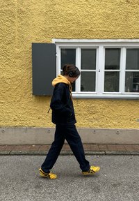 Persoon in gele sneakers, marineblauwe broek en een jack met gele capuchon die langs een gele textuurmuur met een wit raam en een grijze luik loopt.