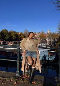 Beige trenchcoat over een lichte trui en denim shorts, gecombineerd met zwarte kniehoge rubberlaarzen. Op de achtergrond een gracht en herfstbomen.