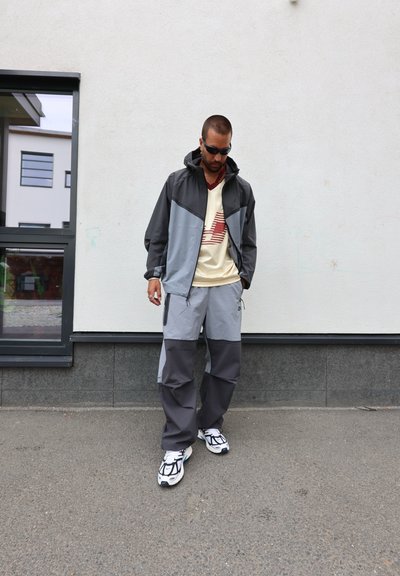 Chaqueta deportiva gris y negra con capucha, combinada con pantalones de chándal grises con acentos más oscuros. La parte superior tiene un diseño gráfico de color claro.