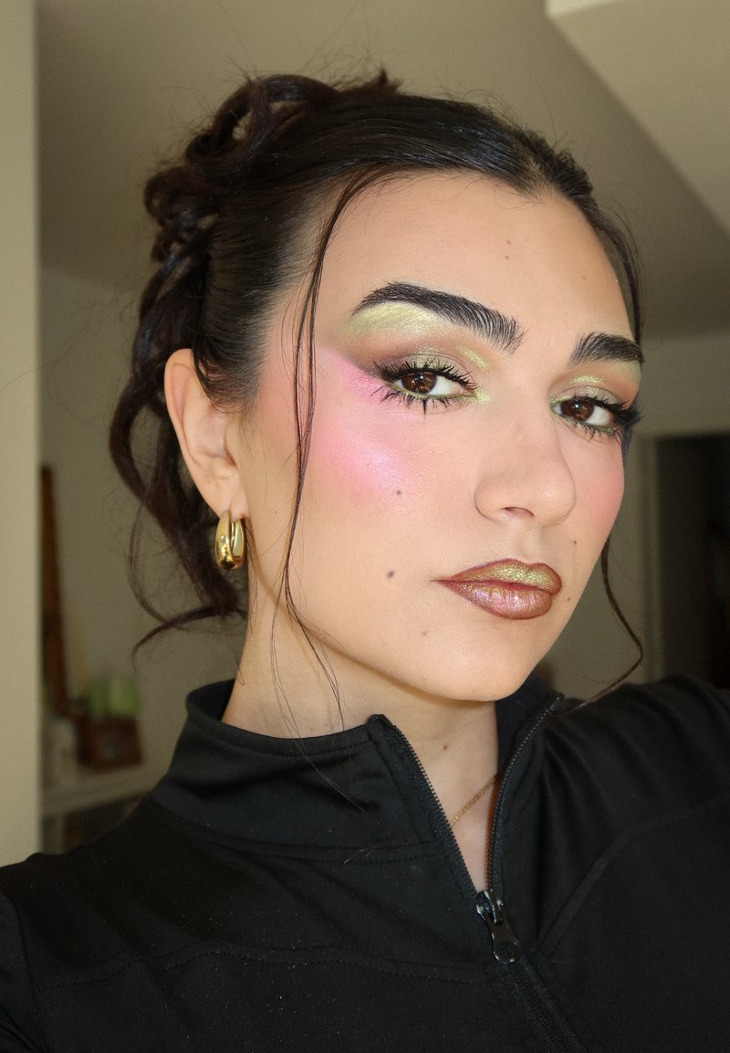 Jeune femme aux cheveux foncés en chignon, portant des boucles d'oreilles créoles dorées, un fard à paupières métallique vert et rose, et un rouge à lèvres bronze, vêtue d'un haut noir zippé.