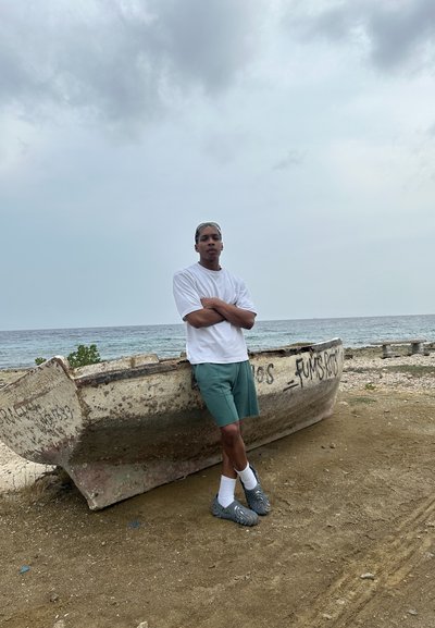 Un barco desgastado y blanco en una playa, con una persona vestida con una camisa blanca y pantalones cortos verdes de pie cerca. Zapatillas grises y un cielo nublado arriba.