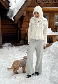 Elefántcsontszínű bolyhos dzseki geometriai mintákkal, világosszürke, testhezálló nadrág és fekete cipő. Egy szürke kabátos kutya áll a hóval borított talajon a közelben.