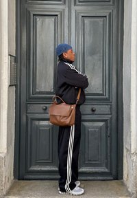 Barna bőr válltáska elöl zsebes, széles pánttal és finoman fémes kiegészítőkkel. Egy fekete melegítőruhát viselő személy hordja, amelyen fehér csíkok vannak.