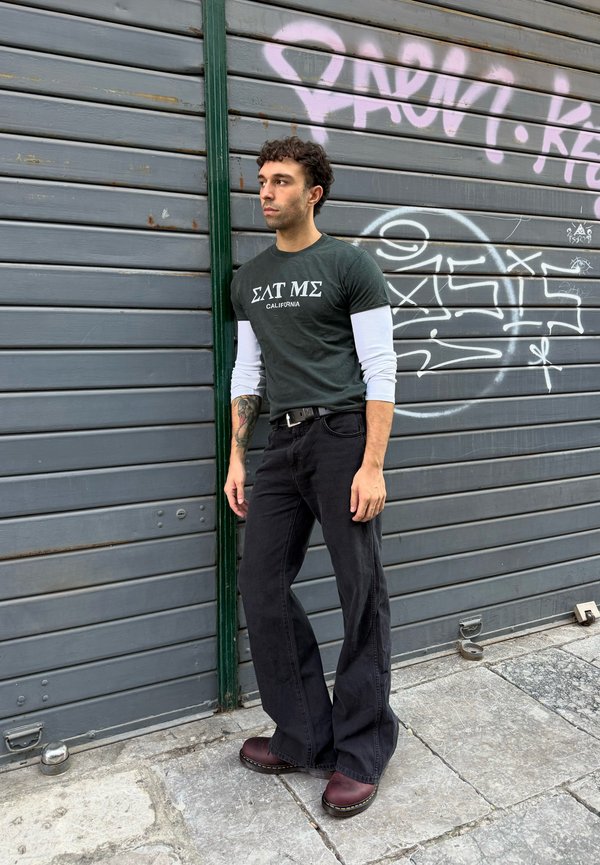 Hombre con tatuaje en el antebrazo está de pie junto a una pared metálica gris con grafiti, vistiendo una camisa verde sobre una camiseta blanca de manga larga y pantalones negros acampanados.