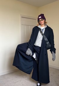 Long manteau bleu marine, pull gris, pantalon bleu marine, sac bandoulière noir, gants gris, bonnet rayé et chaussures à pois avec chaussettes blanches.