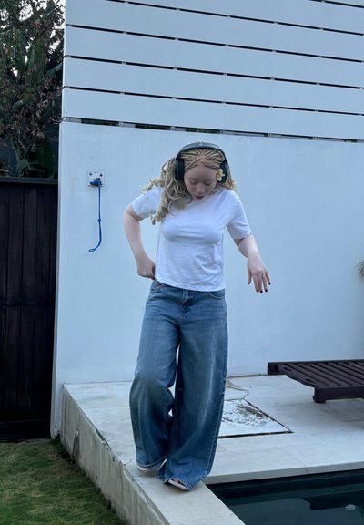 Camiseta blanca y jeans anchos azules usados por una persona. Cabello largo y texturizado con una flor. De pie en un borde de piedra cerca de una piscina.