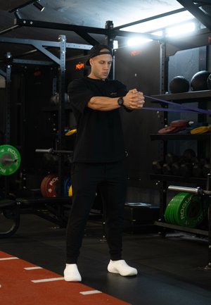 Camiseta y pantalones deportivos negros, zapatos blancos, usando una banda de resistencia morada en un gimnasio con varias pesas y equipos en el fondo.