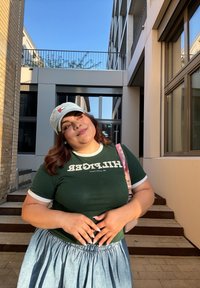 Πράσινο μπλουζάκι με άσπρες λεπτομέρειες που φέρει τη λέξη "HILFIGER". Μπλε τζιν φούστα με μαζεμένο μέσο. Ανοιχτό γκρι καπέλο με λογότυπο. Υπαίθριος χώρος.